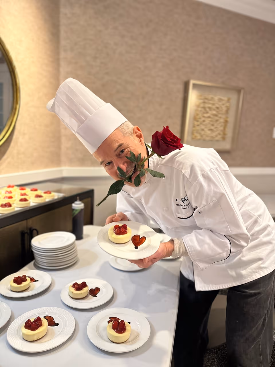 A chef wearing a white chef's hat and coat holds a plate with a small cheesecake topped with strawberries and a heart-shaped decoration. The chef has a red rose held in his mouth and is smiling playfully. Several similar plates with cheesecakes and heart decorations are arranged on a table nearby in a well-lit room with beige walls and framed artwork.