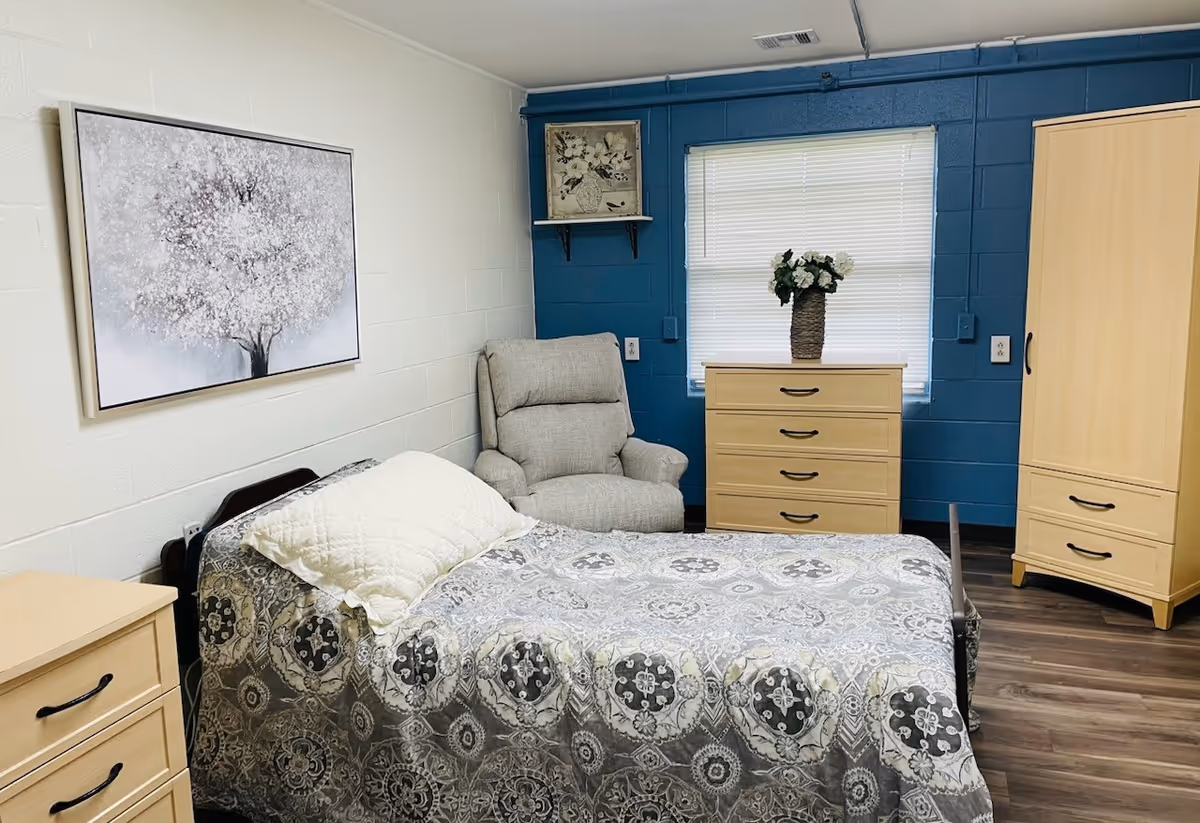 A neatly arranged bedroom in a nursing center featuring a single bed with a patterned bedspread and a white pillow. There is a comfortable gray armchair next to a wooden chest of drawers with a vase of flowers on top. The walls are painted white and blue, with a window covered by blinds and a framed picture of a tree hanging on the wall.