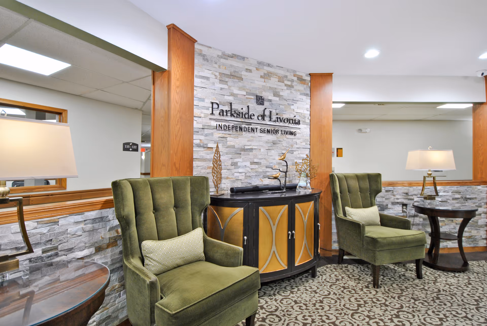 A cozy seating area inside Parkside of Livonia independent senior living facility featuring two green upholstered armchairs with decorative pillows, a patterned carpet, a black and gold cabinet with decorative items on top, and two table lamps on round wooden side tables. The background wall is decorated with stone tiles and the facility's name and tagline.