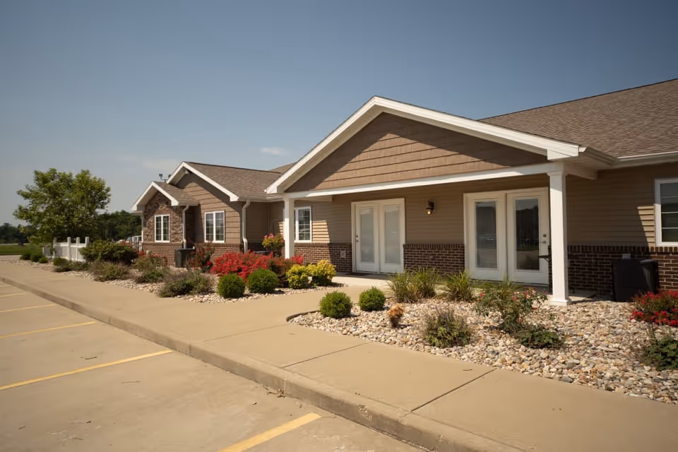 Single-story senior living villa exterior with landscaped rock beds, shrubs, and a walkway leading to white-framed double doors.