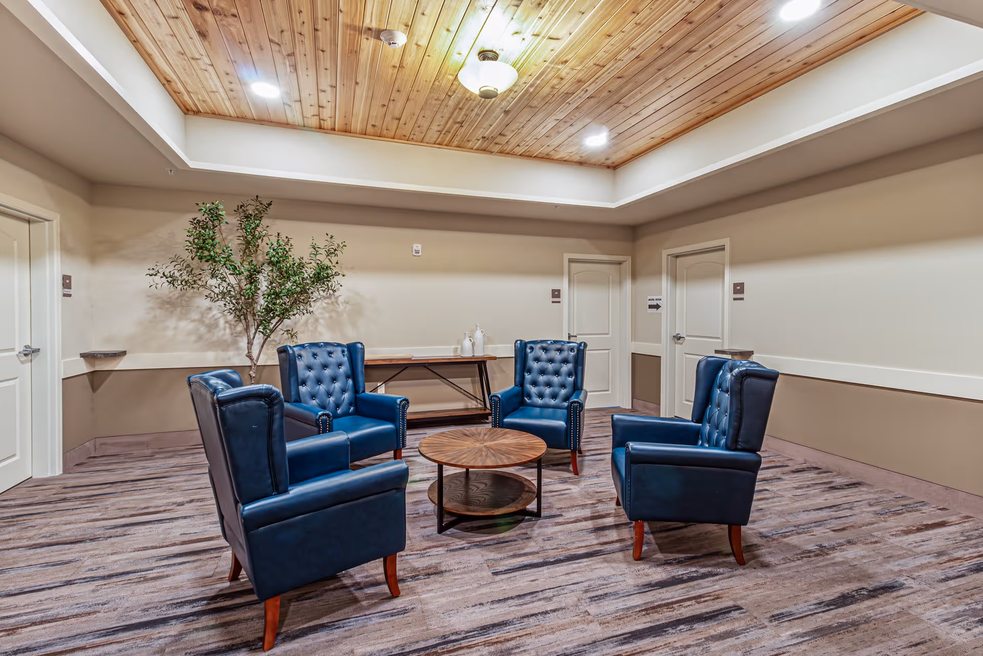 A cozy seating area with four blue leather armchairs arranged around a round wooden coffee table in a room with a wood-paneled ceiling and beige walls. There is a small table with decorative items and a potted plant against the wall.