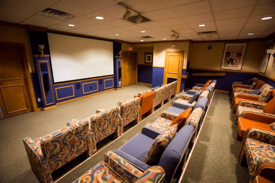 A cozy home theater room with multiple rows of cushioned armchairs and sofas facing a large blank projection screen. The room has warm lighting, wood paneling, and framed posters on the walls.