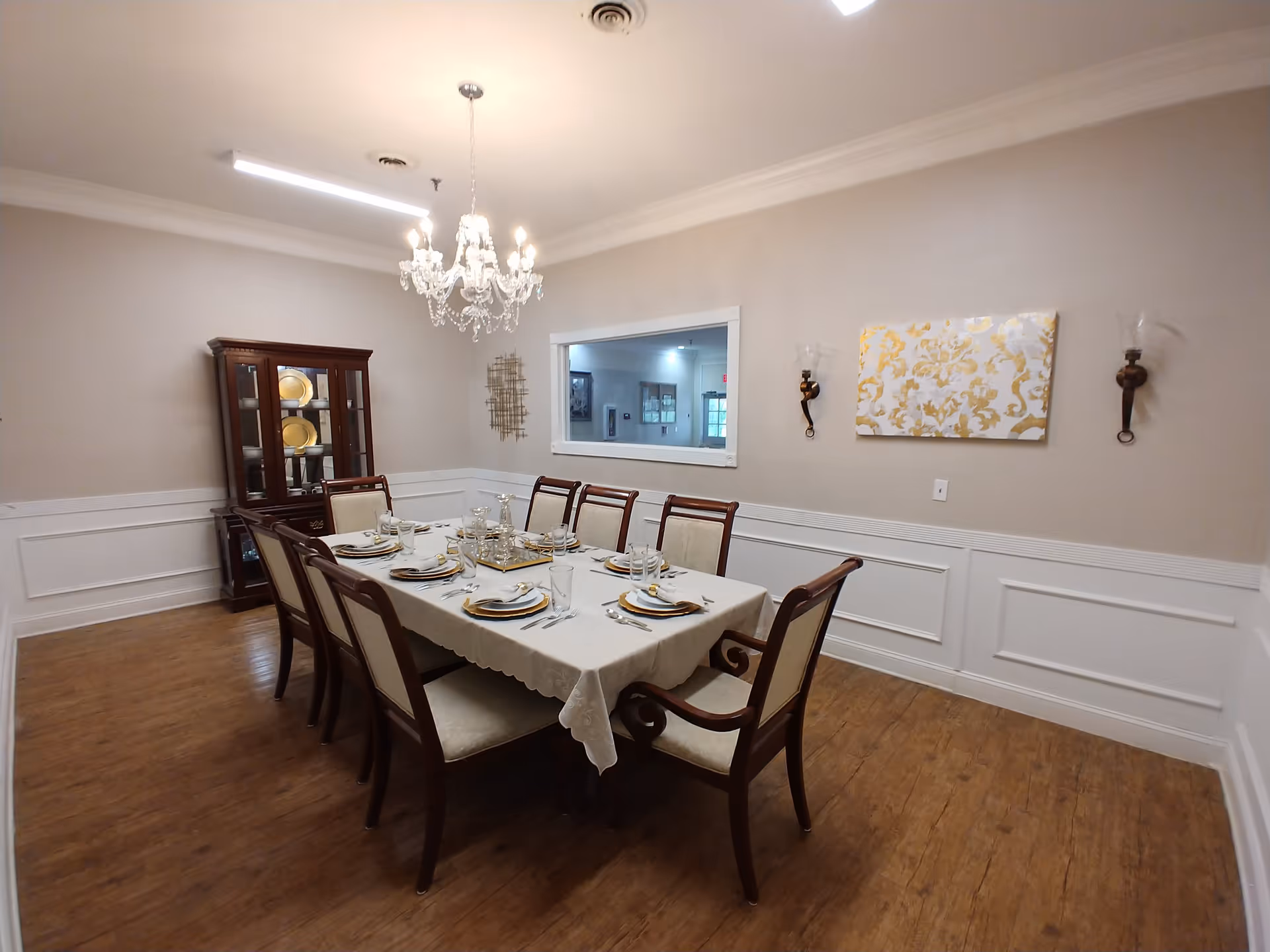 A formal dining room with a rectangular table covered with a white tablecloth set for eight people. The table is surrounded by wooden chairs with cream-colored cushions. A chandelier hangs above the table. There is a wooden china cabinet with gold plates displayed inside against the wall. The walls are beige with white wainscoting, decorated with a gold and white patterned canvas and two wall sconces.