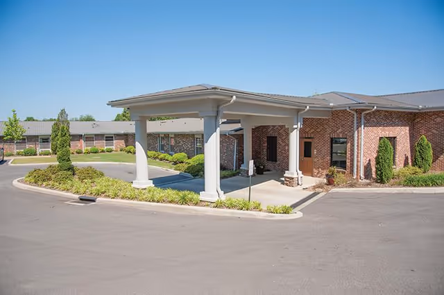 Exterior view of a single-story brick building with a covered entrance supported by white columns, surrounded by landscaped greenery and a paved driveway under a clear blue sky.