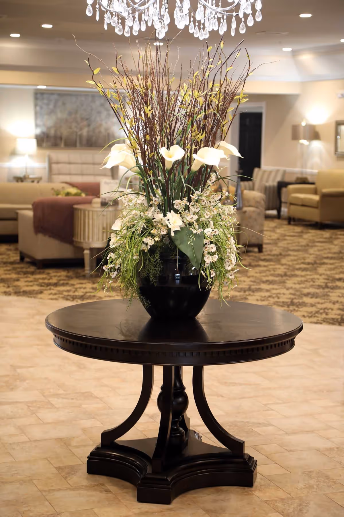 A large black round table with an elaborate floral arrangement in a black vase sits in the center of a well-lit room. The background shows a cozy seating area with beige and brown sofas, armchairs, lamps, and a patterned carpet under soft ceiling lights.