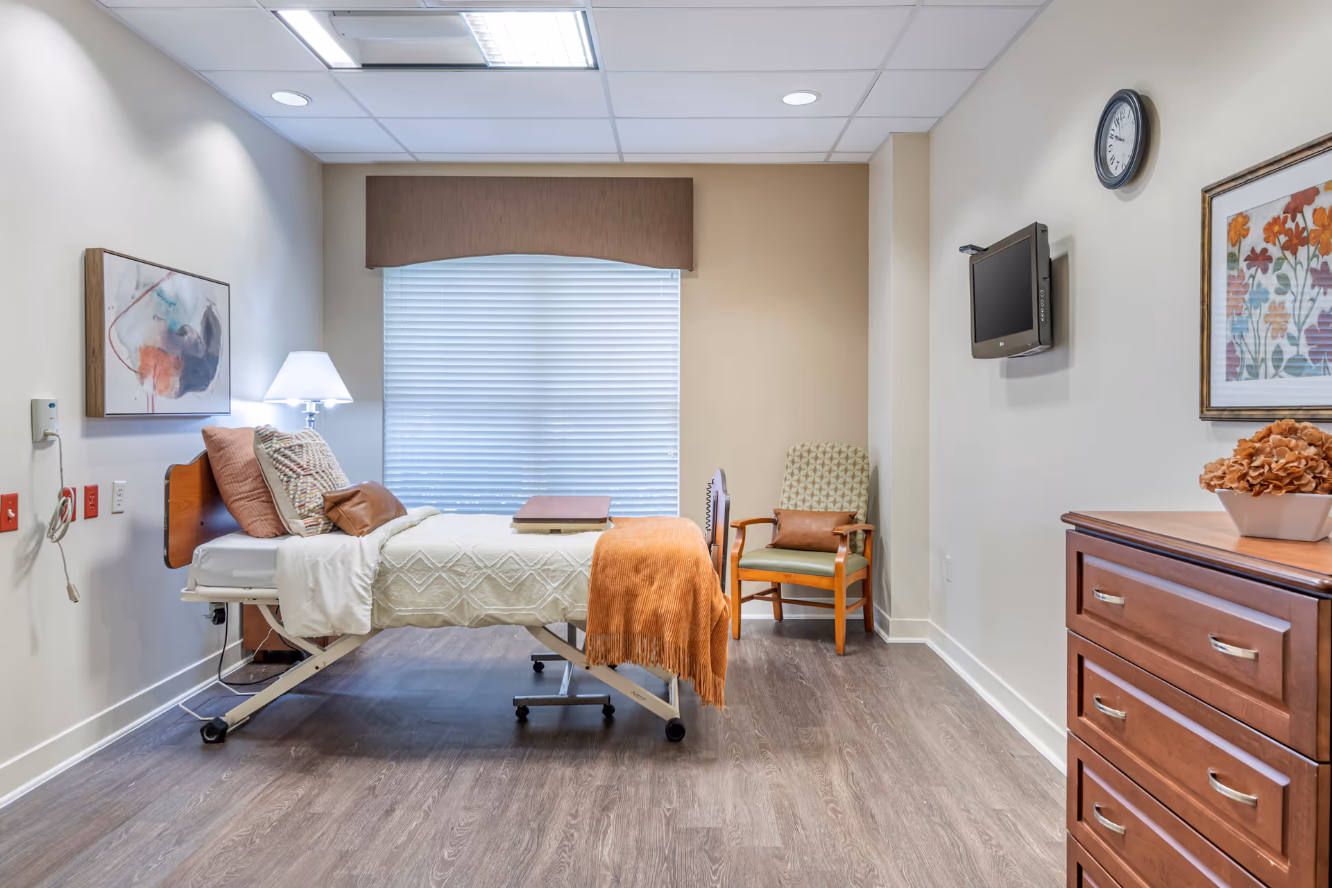 A clean and well-lit healthcare facility room with a hospital bed covered in a beige blanket and orange throw, a cushioned chair with a brown pillow, a wall-mounted TV, a clock, a wooden dresser with a flower arrangement, and a window with closed blinds and a brown valance.