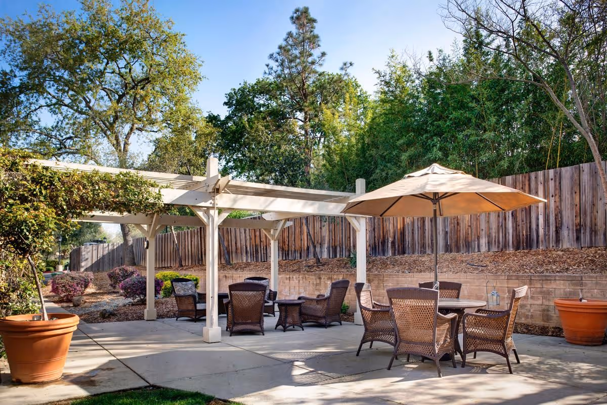 Outdoor patio area with a white pergola and several wicker chairs and tables. One table has a large beige umbrella. The area is surrounded by a wooden fence and trees, with potted plants on the concrete patio.