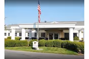 Front exterior view of Sullivan Park Care Center building with white columns, an American flag on a flagpole, and neatly trimmed bushes in front.