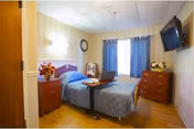 A small bedroom with a single bed covered in a blue quilt, a wooden nightstand with a vase of flowers, a wall-mounted TV, a wooden dresser with a flower arrangement, and a window with blue curtains.