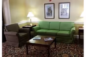 A cozy living room area featuring a green sofa, a patterned armchair, a wooden coffee table with magazines on it, two side tables each with a lamp, and two framed pictures hanging on a light yellow wall above the sofa. The floor is covered with a patterned carpet.