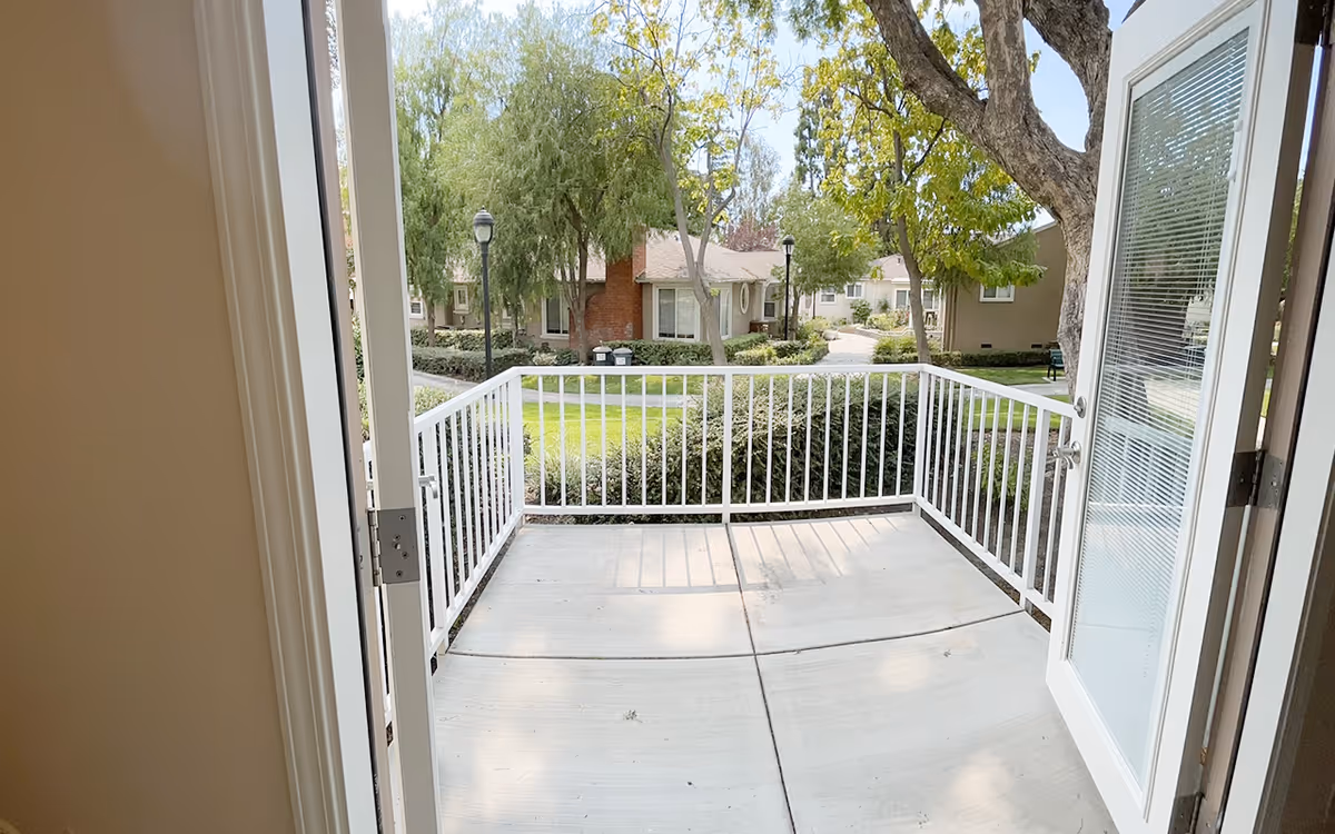 View from inside a room looking out through open double glass doors onto a small concrete patio with white metal railing. Beyond the patio, there are green bushes, trees, and a pathway leading to single-story residential buildings with beige walls and brown roofs.