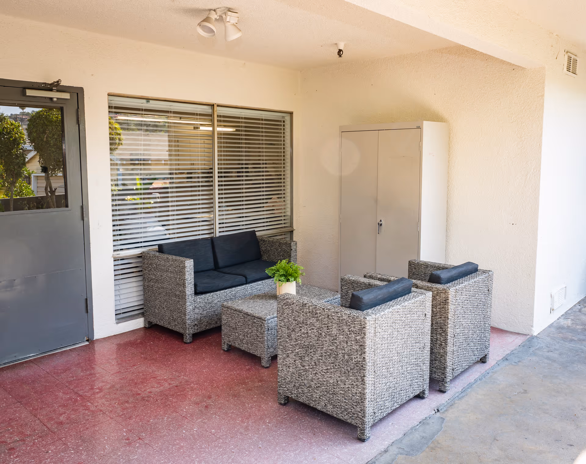 Outdoor covered seating area with a gray wicker loveseat, two matching armchairs with dark cushions, and a small table with a potted plant. There is a gray metal door on the left and a gray storage cabinet against the wall on the right. The floor is a mix of red and gray surfaces.