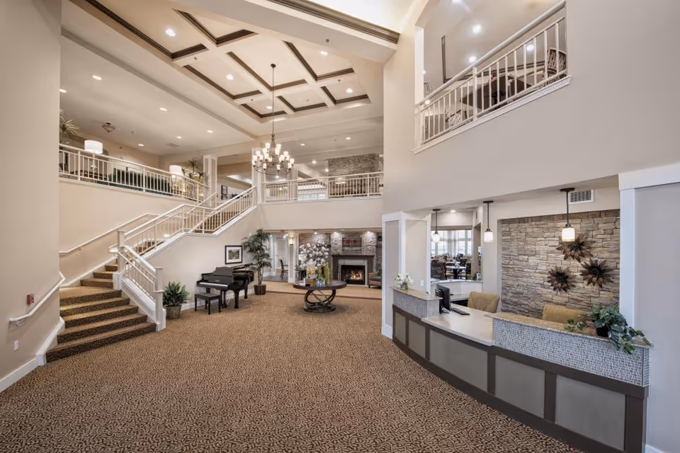 Spacious senior living facility lobby with a carpeted floor, a grand staircase leading to an upper level, a black grand piano with a bench, a round table with floral arrangements, a stone fireplace, and a reception desk with two chairs. The ceiling features recessed lighting and a chandelier, and there are decorative plants and wall art throughout the area.