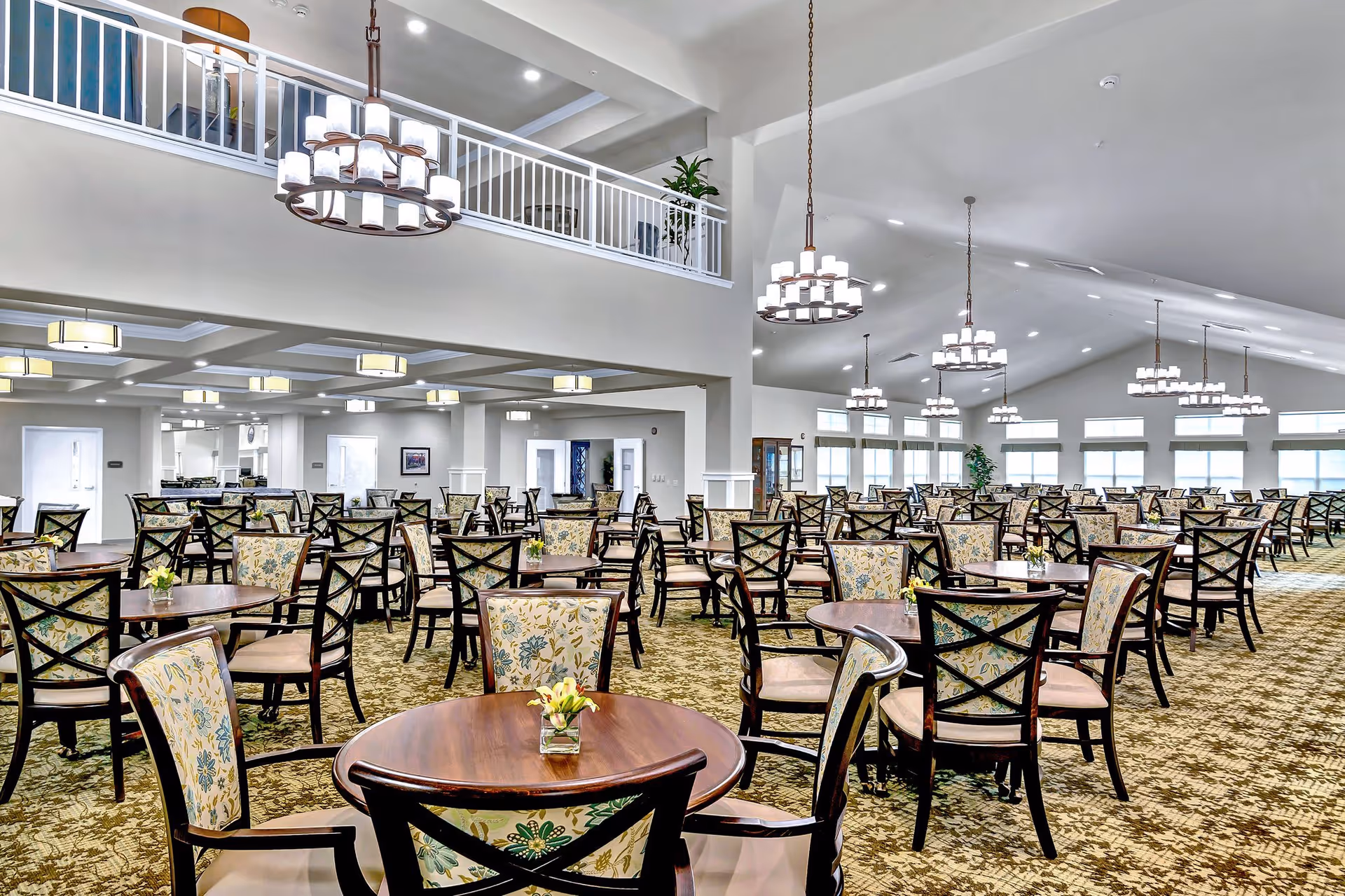 A spacious dining room with multiple round wooden tables and floral upholstered chairs arranged neatly. The room features high ceilings with modern chandeliers hanging down, large windows allowing natural light, and a patterned carpet floor. There is a second-floor balcony with white railings overlooking the dining area.