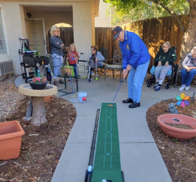 An elderly man playing mini golf on a putting green mat outdoors at a retirement community. Several people, including two elderly women sitting and two children standing, watch him. The setting is a shaded patio area with trees, potted plants, and garden decorations.