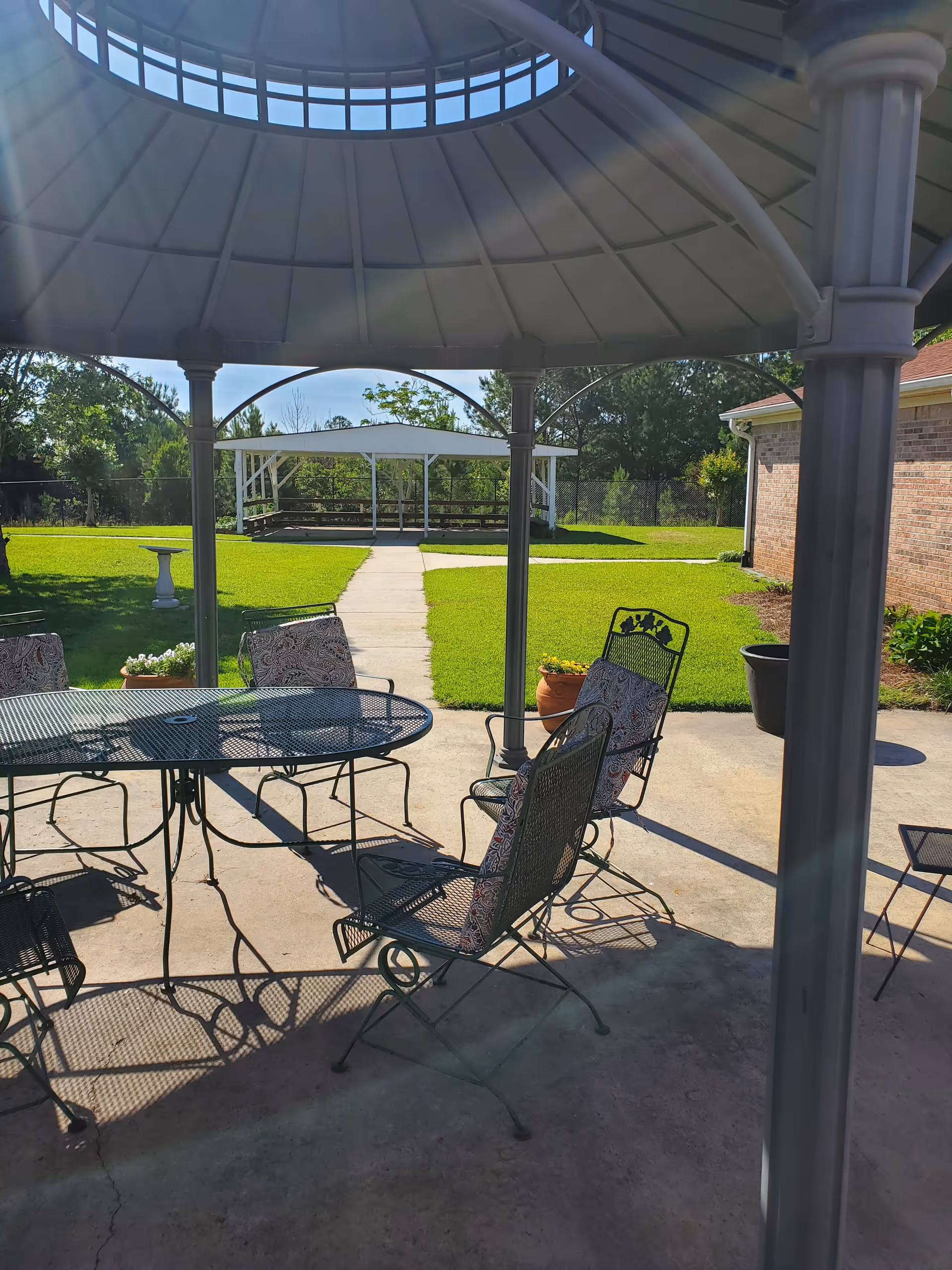 Sunlit outdoor courtyard showing a metal patio table and cushioned chairs under a gazebo with a walkway leading to a white pavilion.