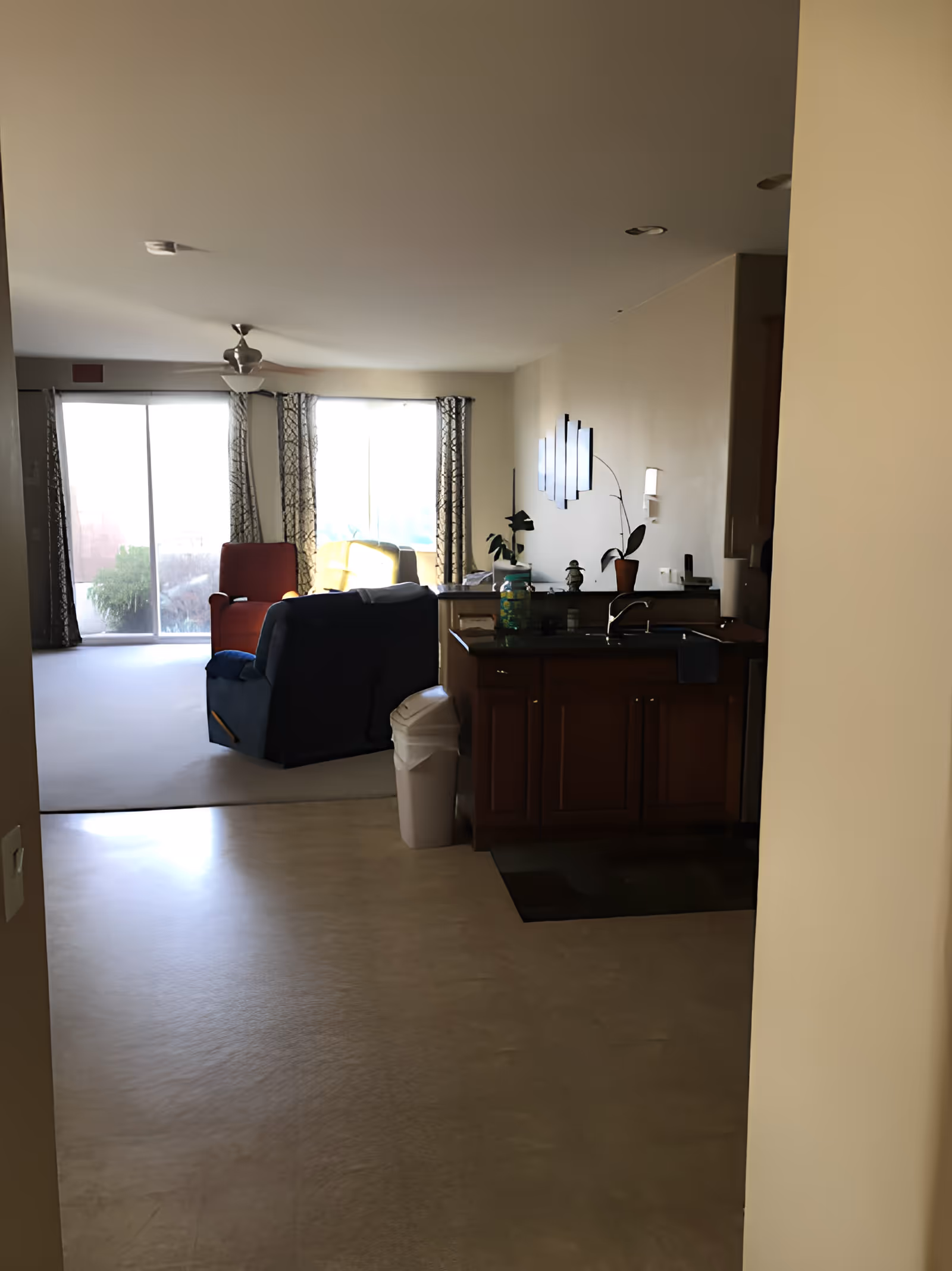 View of a living room and kitchen area in a senior living facility. The living room has a red armchair, a blue recliner, and a yellow chair near large sliding glass doors with patterned curtains. The kitchen area has wooden cabinets, a sink, and a trash can. The walls are light-colored, and there is a ceiling fan in the living room.