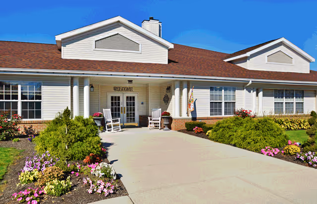 Front exterior view of a single-story senior living facility building with a welcoming entrance featuring two white rocking chairs, a 'WELCOME' sign above the door, and well-maintained flower beds and shrubs along the walkway under a clear blue sky.