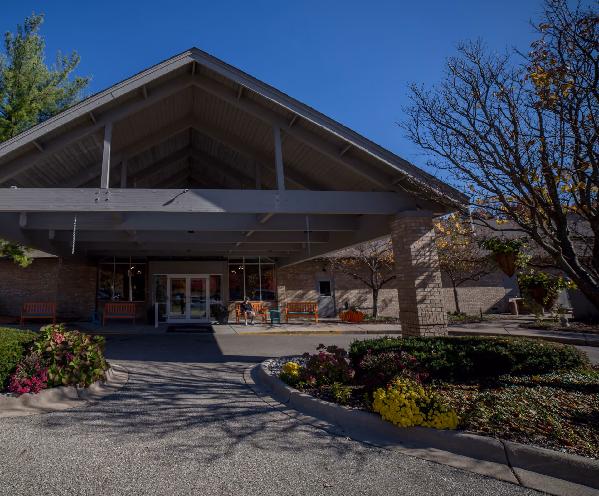 Entrance of a retirement community building with a large covered driveway, benches, and landscaped flower beds. A person is sitting on one of the benches near the entrance. Trees with autumn leaves surround the area under a clear blue sky.