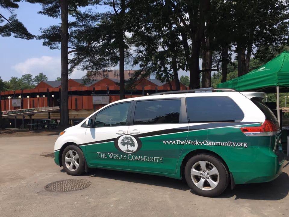 A green and white minivan parked outdoors with The Wesley Community logo and website URL on its side. The background shows trees, a green canopy tent, and a red building structure.