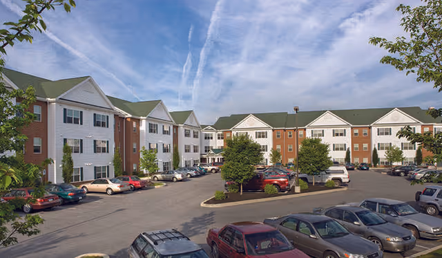 Exterior view of a large senior living facility named Stony Brook Gardens with multiple connected buildings featuring white siding and brick accents, green roofs, and several windows. There is a parking lot in front with numerous parked cars and some trees and shrubs around the area under a partly cloudy sky.