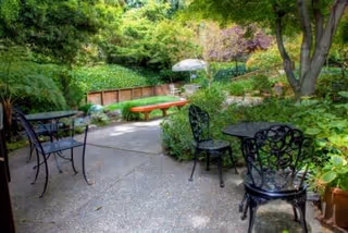 Shaded patio with metal bistro tables and chairs overlooking a lush garden with trees, shrubs, and a path.