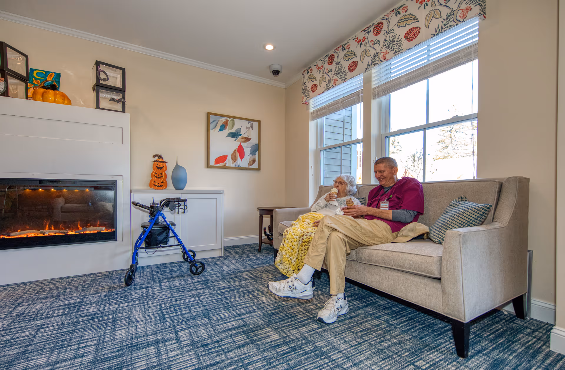 An elderly resident and a staff member sit on a couch in a sunny common living room with a fireplace, walker, and large windows.