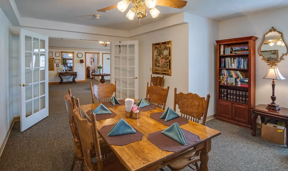 A dining room in a senior living facility with a wooden table set for six people with folded blue napkins on brown placemats. The room has wooden chairs, a ceiling fan with lights, a bookshelf filled with books, a small table with a lamp and a decorative mirror on the wall. French doors open to another room with additional furniture and wall decorations.