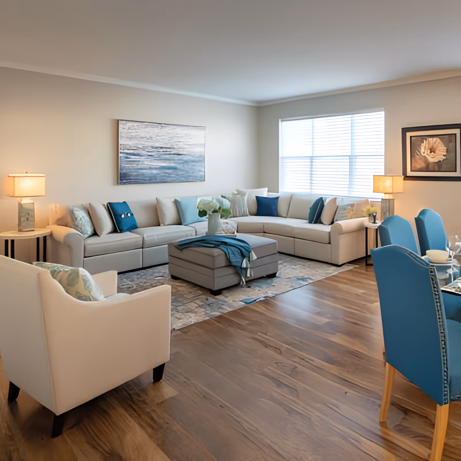 A bright and spacious living room with a large beige sectional sofa adorned with blue and white cushions, a matching ottoman with a blue throw, a white armchair, two side tables with lamps, a large window with blinds, and a dining area with blue upholstered chairs and a glass table. The room features wooden flooring and neutral-colored walls with framed artwork.
