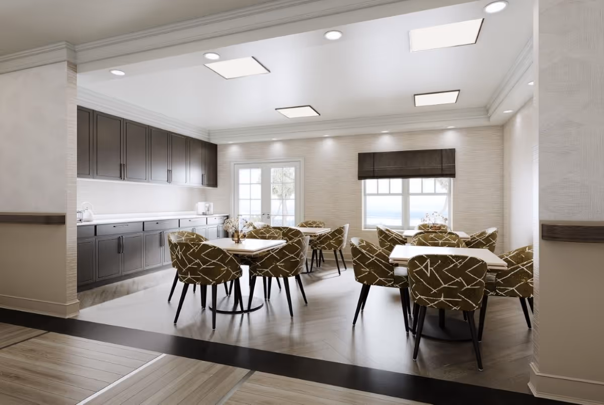 Bright communal dining room with multiple tables and patterned chairs, cabinetry along one wall, and windows letting in natural light.