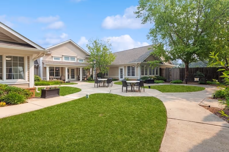 Outdoor courtyard area of HeartFields Assisted Living at Frederick featuring green grass, paved walkways, several round tables with chairs, trees, and beige buildings with white trim under a blue sky with some clouds.