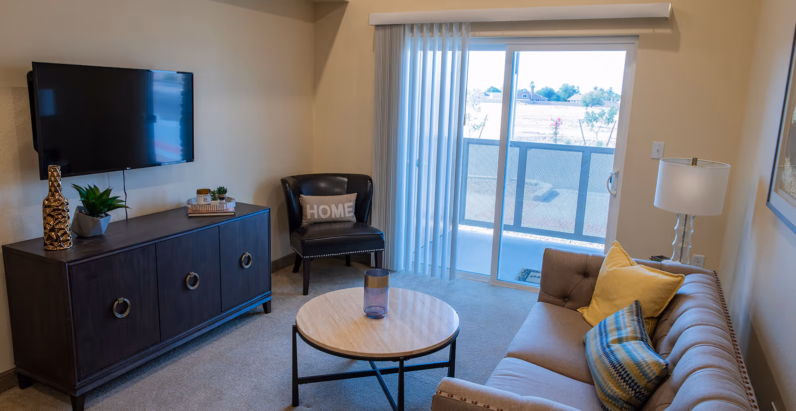 A cozy living room with a beige tufted sofa adorned with yellow and patterned pillows, a round wooden coffee table with a glass vase, a black leather chair with a pillow that says 'HOME', a dark wooden TV stand with decorative items, a wall-mounted flat-screen TV, a floor lamp, and sliding glass doors with vertical blinds leading to an outdoor area.