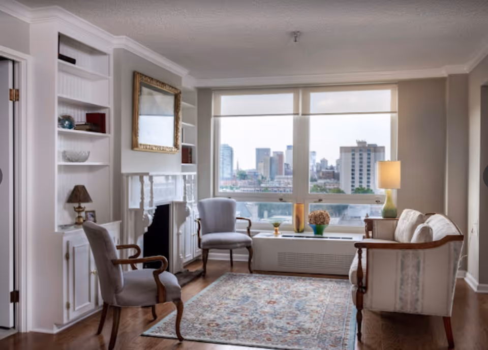 A cozy living room with two upholstered armchairs and a sofa arranged around a patterned area rug. There is a white fireplace with a gold-framed mirror above it, built-in white shelves with decorative items, and a large window offering a cityscape view. A table lamp is placed on a side table next to the sofa.