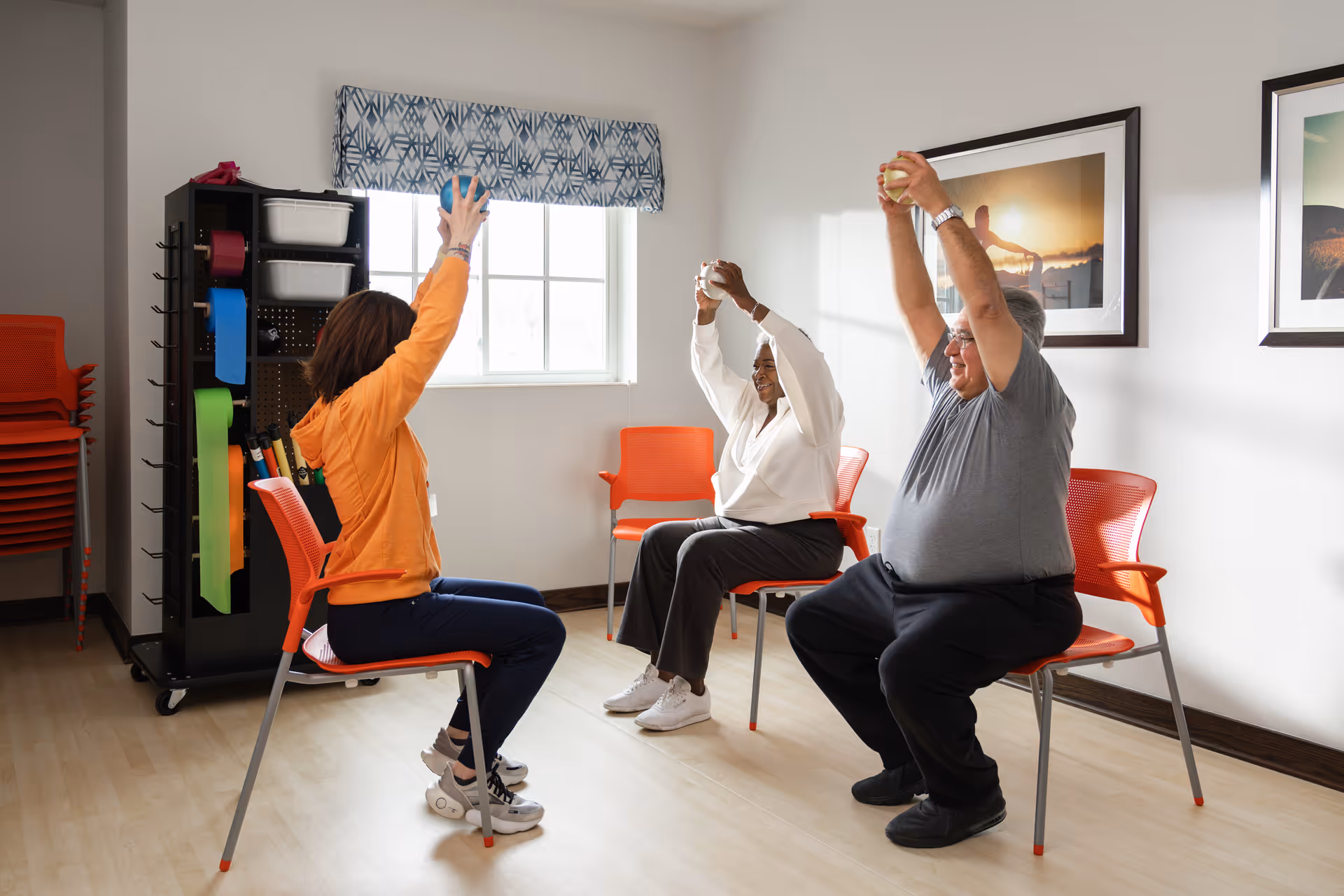 Three adults sitting on orange chairs in a bright room, participating in a seated exercise by raising their arms above their heads while holding small balls. The room has light-colored walls, a window with a patterned valance, and framed pictures on the wall.