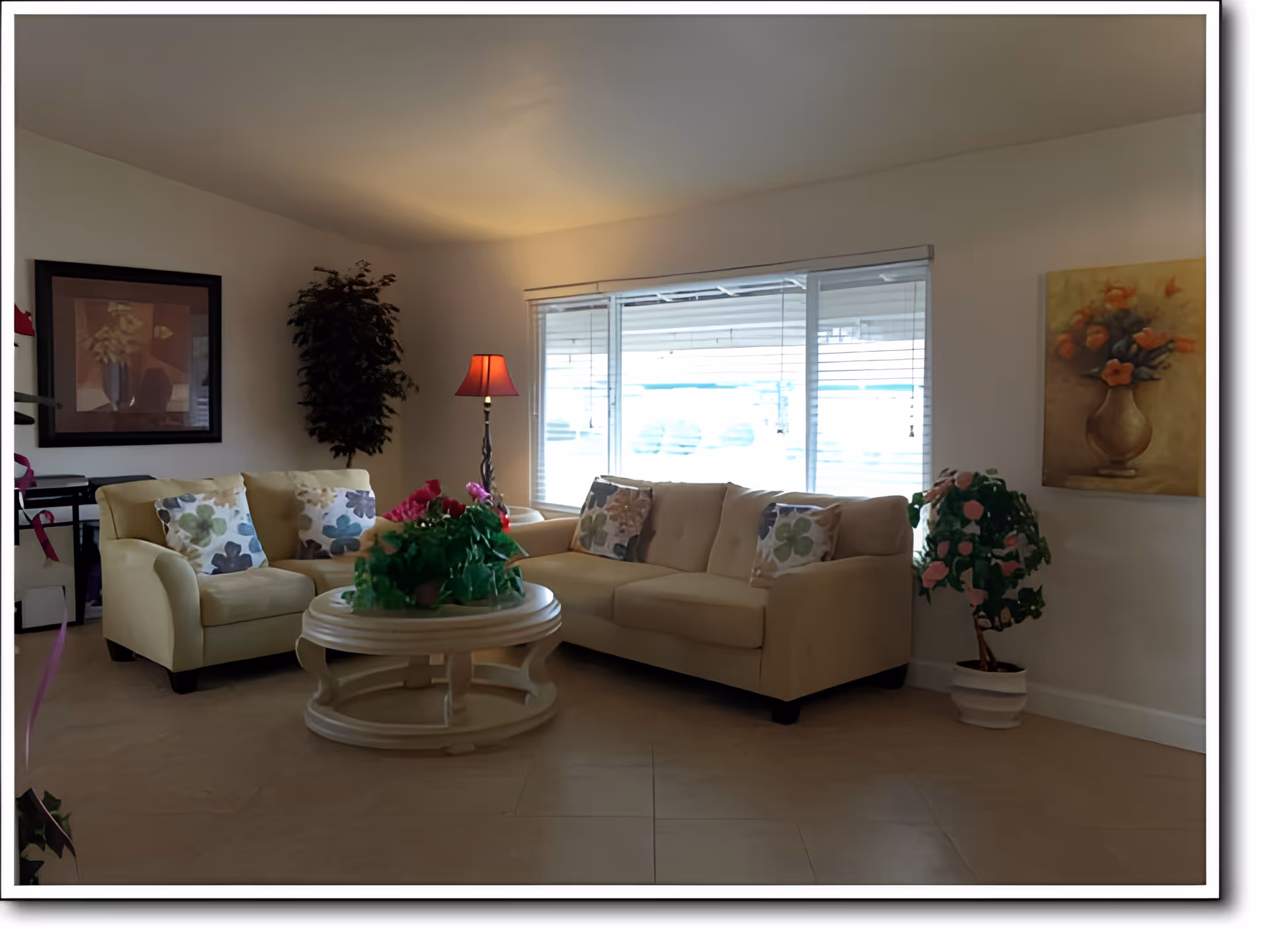 A cozy living room with beige sofas adorned with floral cushions, a round white coffee table with a green plant centerpiece, a floor lamp with a red shade, two potted plants, and framed floral artwork on the walls. Large windows with blinds allow natural light into the room.