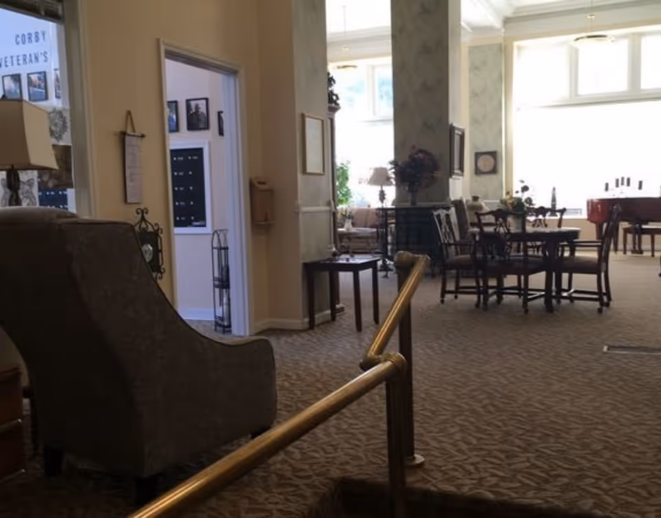Interior view of a senior living facility common area with carpeted floors, a comfortable armchair, a small table, and a dining table with chairs. There is a piano in the background near large windows letting in natural light. A brass handrail is visible along a small step down into the room.