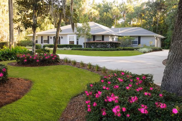 A single-story residential building surrounded by a well-maintained garden with green grass, pink flowering bushes, and tall trees in the background. A curved driveway leads up to the building.