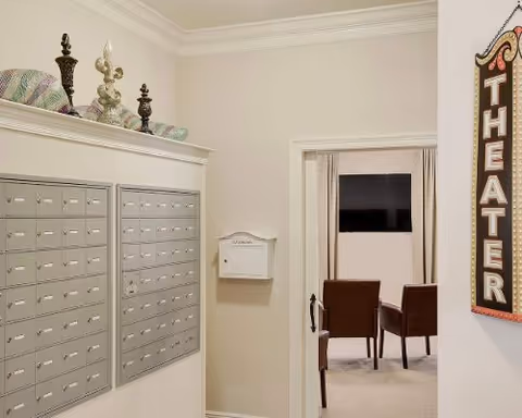 Interior view of a senior living facility hallway with multiple mailboxes mounted on the wall, a white mailbox labeled 'VILLAGE', decorative items on a ledge above the mailboxes, and a doorway leading to a small theater room with chairs and a TV screen. A vertical sign on the wall reads 'THEATER'.