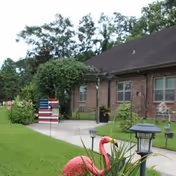 Front exterior of a brick senior living building with a grassy lawn, a pink flamingo lawn ornament, a pathway, and an American flag-themed yard decoration.