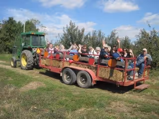 A group of people sitting on a hayride trailer being pulled by a green tractor on a grassy path with trees and a blue sky in the background. The people are waving and appear to be enjoying the outdoor ride.