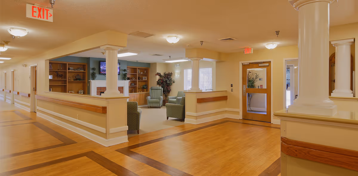 Well-lit interior common area of a memory care facility with seating around a fireplace, built-in shelves, columns, and a corridor with exit signs.