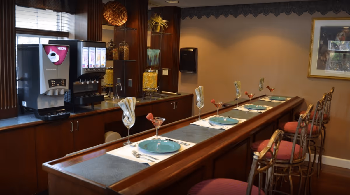 A dining area with a long wooden counter set with blue plates, silverware, and glasses. Behind the counter, there is a beverage dispenser and a cereal dispenser on a countertop with cabinets below. Several high chairs with red cushions are placed along the counter. The walls are beige with a framed picture hanging on one side.