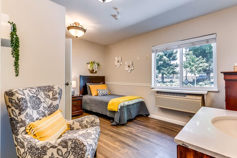 A cozy bedroom with a single bed covered in a gray bedspread and yellow throw blanket, a wooden nightstand with a lamp, a floral-patterned armchair with a yellow pillow, a window with a view of trees outside, and a wall-mounted air conditioning unit below the window. The room has wooden flooring, beige walls, and decorative wall flowers above the bed.