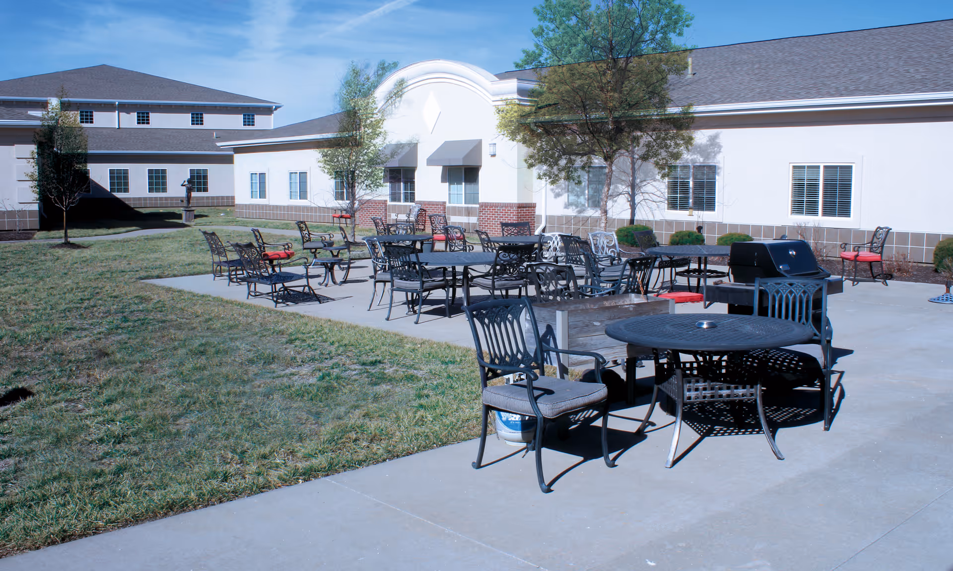 Outdoor patio area at Pegasus Landing of Overland Park with multiple metal tables and chairs arranged on a concrete surface, a barbecue grill, and a grassy lawn surrounding the patio. The building exterior is visible in the background with windows and some small trees.