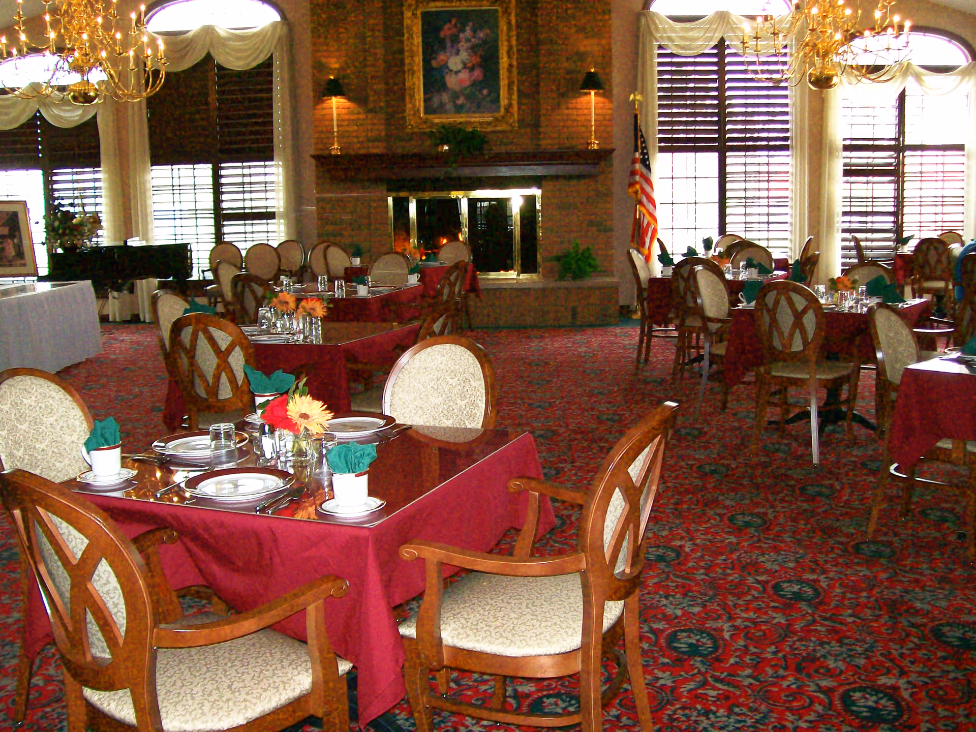 A dining room with multiple tables covered in burgundy tablecloths, set with plates, cups, and green napkins. The room features upholstered wooden chairs, a large brick fireplace with a floral painting above it, chandeliers, and large windows with wooden blinds and white drapes. An American flag is visible near the fireplace.