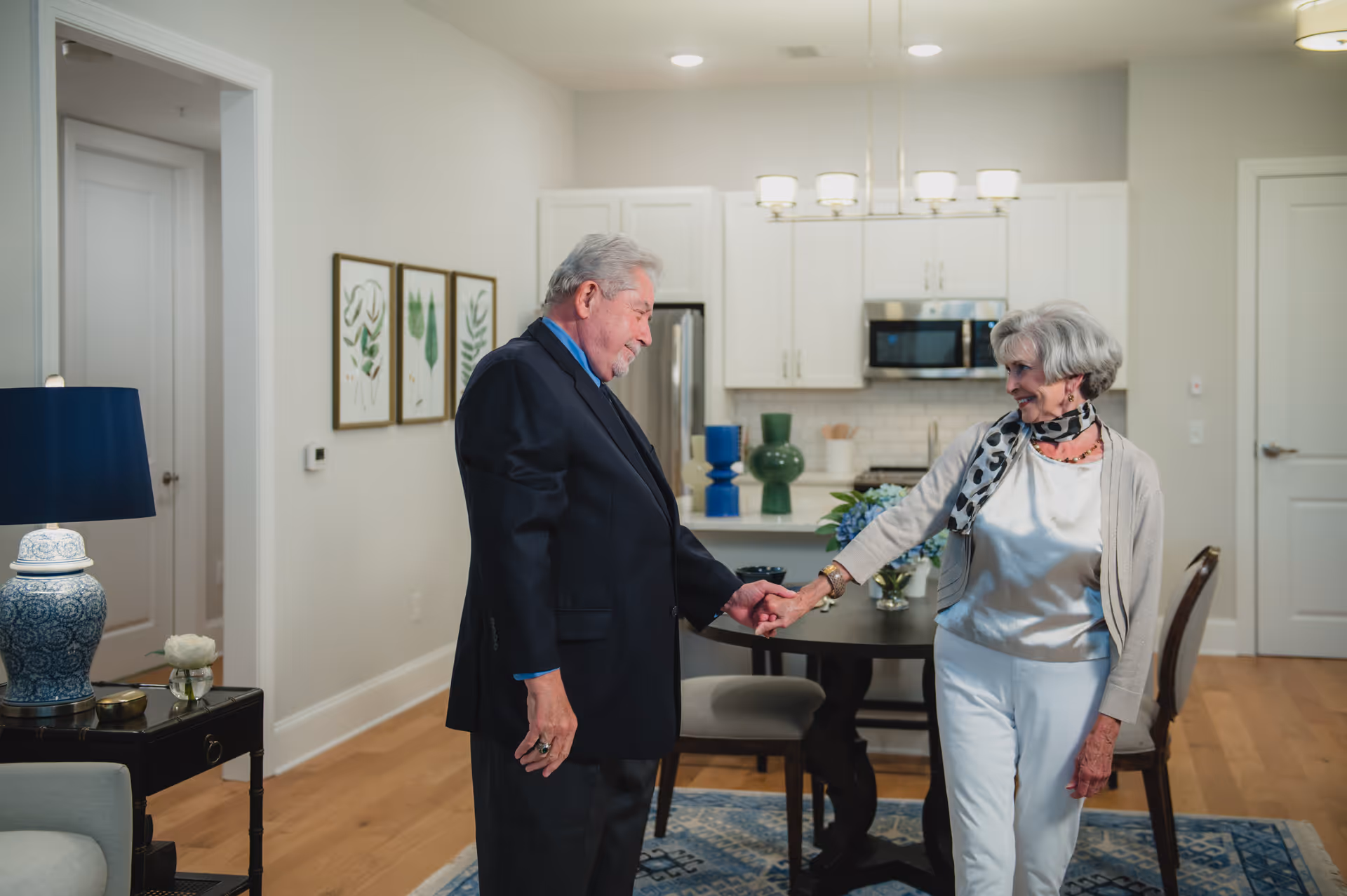 An elderly man and woman holding hands and smiling at each other in a modern kitchen and dining area. The kitchen has white cabinets, stainless steel appliances, and a round dining table with chairs. The room is decorated with framed botanical prints on the wall and a blue patterned rug on the floor.