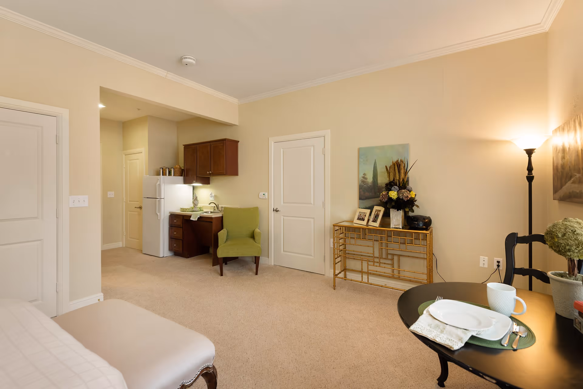 A cozy interior room with beige walls and carpeted floor featuring a small kitchenette with a refrigerator, sink, and wooden cabinets. There is a green upholstered chair next to the kitchenette. A decorative console table with framed pictures and a flower arrangement is against the wall. A round dining table is set with a plate, cup, and utensils, and a floor lamp provides warm lighting.