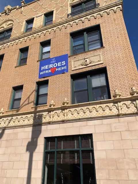 Exterior view of a multi-story brick building with decorative architectural details. A blue sign on the building reads 'Eden HEROES WORK HERE' with a red heart symbol. The building has several windows with dark green frames and a clear blue sky in the background.