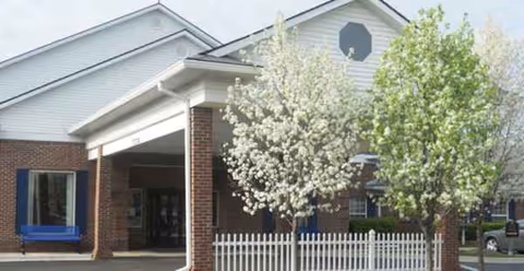 Exterior view of a senior living facility entrance with trees and a white fence.