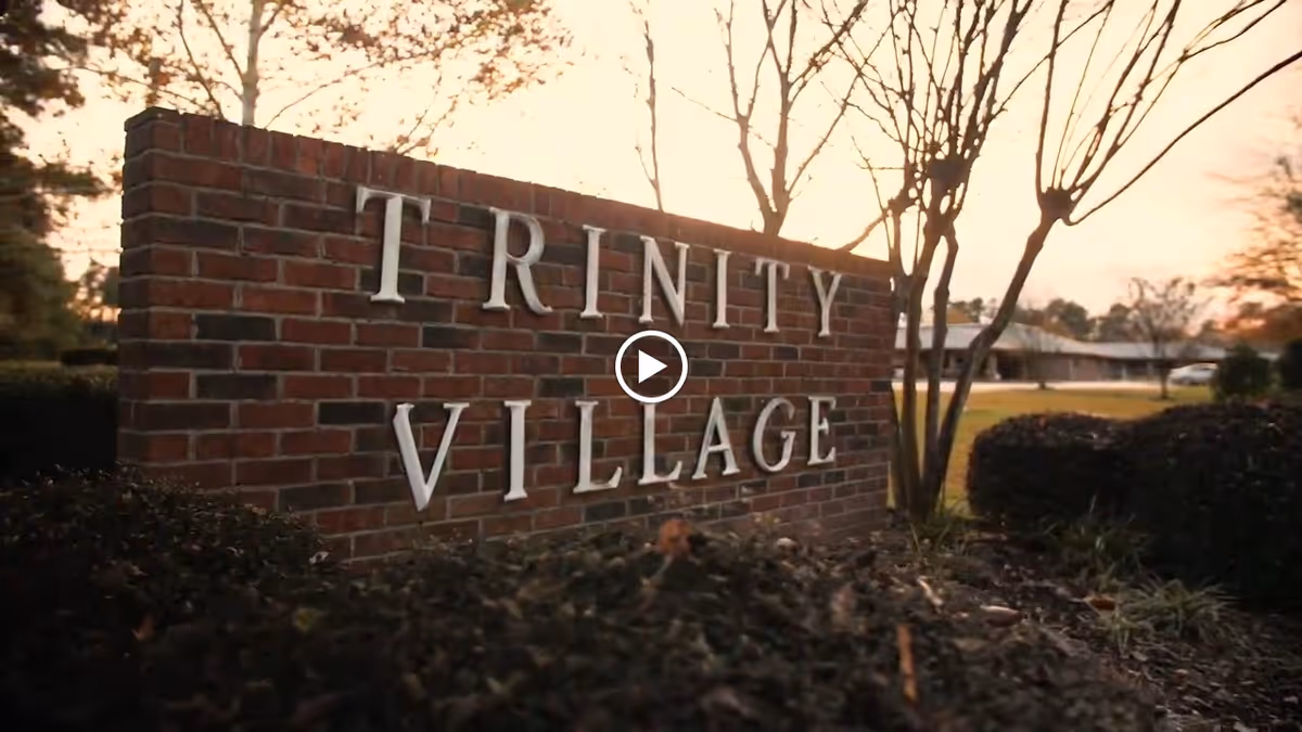 A brick sign with silver letters spelling 'TRINITY VILLAGE' surrounded by bushes and trees, with a building and sunset sky in the background.